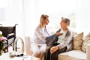 A nurse sitting with an elderly woman. The nurse has a stethescope on the woman's chest.