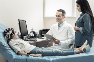 a girl on a bed with an EEG electrode array on her head sitting with smiling doctor in a white lab coat and a woman in a denim jacket, presumably her motherBrain Wave Test for Epilepsy