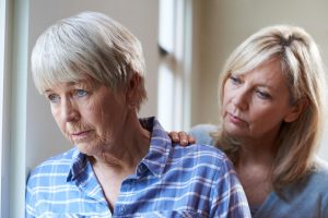 An older woman appearing sad standing next to a younger woman with blond hair. The younger woman has her hand on the older woman's shoulder.