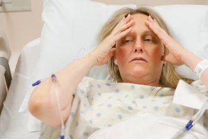 middle age blond woman in a hospital bed and in a white hospital gown with an intravenous line running, holding her hands up to her forehead