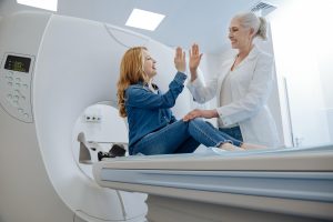 woman with red hair sitting on the platform of an MRI machine "giving five" to the blond woman MRI technician in a white clinical coat