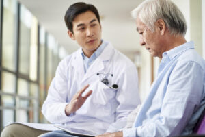 A young doctor in a white clinical coat sits next to a depressed looking elderly gentleman in a light blue dress shirt. They are in a bright open room with large windows.