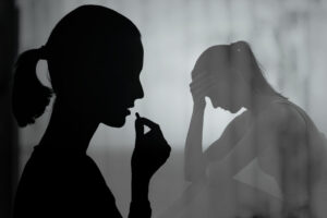 black silhouettes against a grey background of a woman with a ponytail taking a pill and another woman, more distant, with her head in her hand