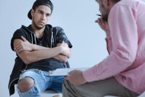 Angry defensive young man in a dark t-shirt and sweatshirt sitting with arms crossed facing a male doctor in a pink sweater