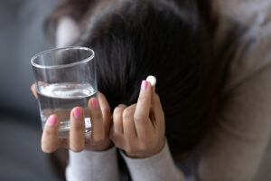 Woman with her head down holding a glass of water in one hand and a white pill in the other