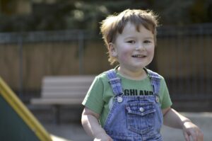 a boy about 5 years old on a playground with a big smile in denim overalls and a green t-shirt