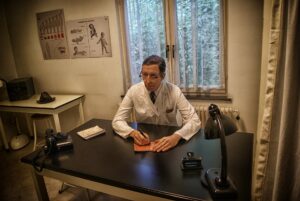 A 1940s physician, wearing a white clinical coat, sits in his office at a simple desk writing on a pad. There's a lamp and a telephone on the desk. Outside the windows behind him are tall evergreen trees with snow.