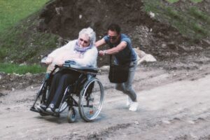 a large elderly gray-haired woman in a wheelchair is pushed up a hill outdoors on a gravel road by a young man exerting a lot of effort to push her up the hill