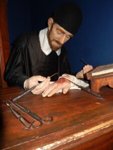 a bearded physician in medieval clothes with a black cap and black gown with a white collar is dissecting a hand on a well-worn wooden clinical work bench.