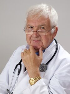 an elderly male physician with white hair, wearing rimless glasses slid down his nose, with a white clinical coat, a stethoscope around his neck, and an expensive gold watch on his wrist