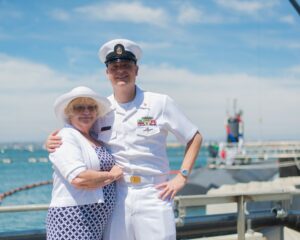 A shorter, older woman is standing with a tall handsome middle-age man in dress white military uniform, his arm around her, both smiling at the camera. A ship and the ocean are behind them. It appears to be an officer in the Navy and his proud mother.