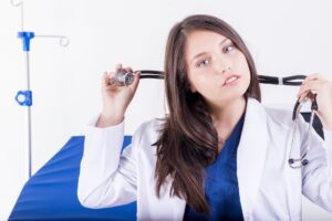 A young attractive brunette woman doctor wearing a white clinical coat and a dark blue blouse is looking at the camera and starting to drape her stethoscope around her neck.