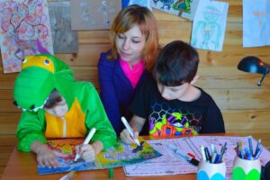 two young boys, one in a green alligator costume, at a desk, coloring in a coloring-book with a kindly teacher watching from behind them