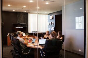 four people (including a guy in a grey tee-shirt and a baseball cap) seen through a clear glass wall sitting around an oval conference table, laptop computers in front of them, inside a comfortable conference room