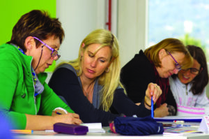 four women in pairs working on and discussing documents along one side of a work table