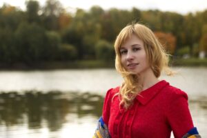 smiling young woman with long blond hair in a red top standing in front of a lake with trees (out of focus) along the far shore