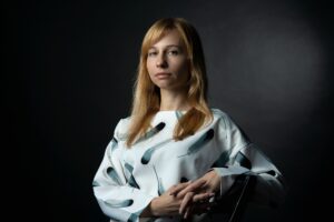 formal posed portrait of a young woman with long blond hair in a white top with a blue-gray feather pattern, a serious expression on her face, against a photographer's grey background