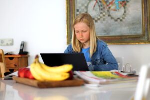 Young blond girl about 10 years old looking studious and sitting at a kitchen table with bananas and peaches in a tray in the foreground. She's looking at a laptop computer and has books beside her on the table.