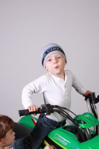Young boy about six years old perched on a Speedee brand bright green all-terrain quad bike with a blank background, perhaps in a studio. He's looking excited and confident, hands on the handlebars and making motor revving sounds while a friend that's his age looks on. On his knitted cap it says, "Be Cool".