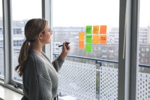 woman in a bulky grey sweater reviewing writing on green and orange Post-It notes posted on a clear window with a distant city view outside the window