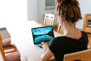 woman with red hair sitting at a wood table with her back to the camera working on a netbook (small) laptop computer with a colorful bluish picture on the screen