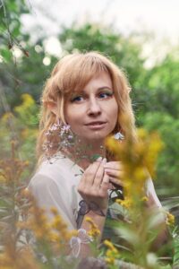 smiling young woman with long blond hair in a white top holding a small flower perhaps in a woods (out of focus) with a clearing of flower