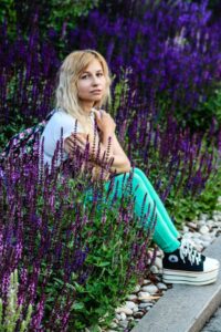 smiling young woman with long blond hair in a light blue top, bright turquois jeans, and black and white Converse athletic shoes, arms crossed in front of her, sitting amongst a garden of tall purple flowers
