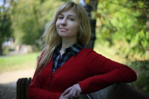 smiling young woman with long blond hair in a red sweater over a dark blue plaid check shirt sitting on a park bench in a park with trees (out of focus) in the background