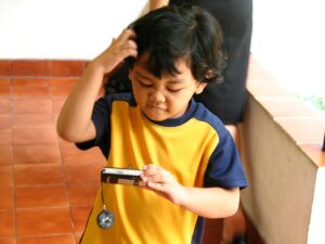 A little boy with black hair wearing a blue-and-yellow top scratches his head as he stares at a little camera in his hand.