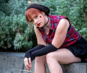 Woman with red hair, red lipstick, and a red heart on her cheek sitting on a gray block wall in front of shrubs in black shorts and a sleeveless check red flannel top staring into the camera with an incredulous look.