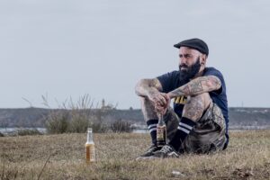 Heavily tattooed Caucasian man with serious look on his face. He has a black beard and is wearing a tee shirt, shorts, sports socks, and a flat cap, sitting in an open field on dry, brown grass with an open bottle of beer dangling from his hands and another open bottle of beer sitting on the grass in front of him.