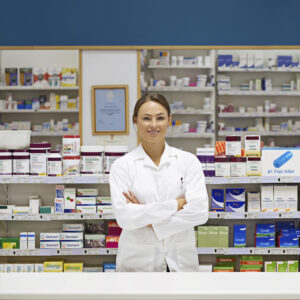 A young attractive woman pharmacist in a white lab coat, smiling and arms folded, stands in the pharmacy with hundreds of packages, vials, and bottles of medications on the shelves behind her