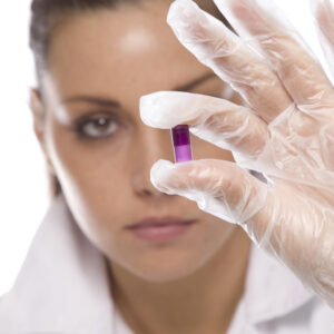 A portrait-style photo of a young, attractive woman pharmaceutical researcher in a white lab coat as she holds up a two-tone purple capsule in a gloved hand in front of her face
