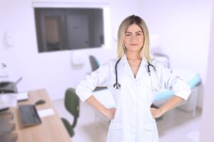 Attractive young woman physician in a form-fitting white clinical coat stands in a medical exam room and faces and smiles at the camera, stethoscope around her neck and hands on her hips.