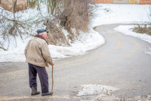 An elderly man in a tan coat and blue cap walks with a cane at the start of a long downhill winding road with snow and bare winter trees on the banks of the road.
