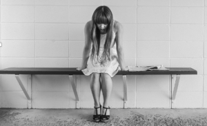 Black and white photo of a young woman in a white dress and black strap shoes sitting alone and looking down at the floor on a bare bench attached to a tile wall. The setting feels cold and stark.
