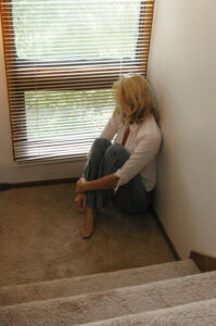 Barefoot blond woman in blue jeans and a white blouse sitting on the floor in a corner on a carpeted staircase landing staring out a venetian-blind-covered window at the green trees outside.