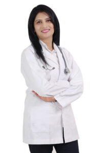 Attractive, tall, smiling woman physician with long, dark hair stands looking at the camera, stethoscope around her neck and wearing a short-style white clinical lab coat, arms loosely crossed.