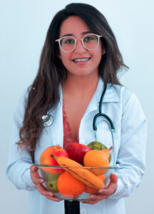 Friendly, smiling woman nutritionist wearing fashionable large white-framed glasses. She has on a white clinical lab coat with a doctor's stethoscope around her neck. She holds an attractive clear glass bowl filled with whole fruits in front of her.